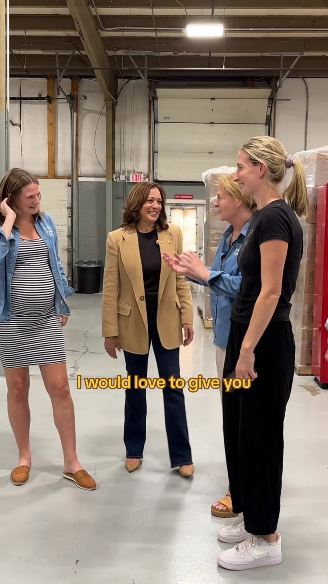 The image features four women standing in a warehouse setting. The woman in the center is wearing a tan blazer and dark pants, and seems to be the focus. The other three women stand around her, two dressed in casual clothes, and one is pregnant. The background shows a loading dock and shelves.