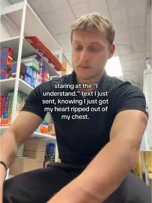A young man, positioned slightly off-center, sits with a somber expression. His head is angled down, eyes looking downward. He wears a black t-shirt, and his arms are visible as he's seated. Behind him, bookshelves with organized items are visible, adding a sense of a well-kept space. The lighting is somewhat dim, focusing on the man. The text overlayed on the image conveys a melancholic tone.