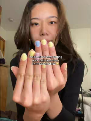 A young woman with long brown hair is holding up her hands, showing off colorful manicured nails. Her hands are in the foreground, with a soft-focused background including a wooden cabinet and bedding. Text is overlaid, expressing a thought about the cost of nail services.