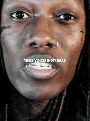 Close-up shot of a person's face with heavily emphasized eyes and mouth. The skin appears weathered with dark, ashy tones and visible textured with subtle shadows, piercings on the nose and lip. Overlaid on the image is the text "THESE VOICES IN MY HEAD" in a bold, white font, strategically positioned to draw the eye.