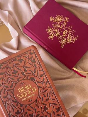 The image displays two closed books resting on a softly draped surface. The book on the right is a deep burgundy color with a gold floral design. The book on the left is a lighter brown color with a circular emblem in the middle, covered in what appears to be an embossed floral pattern.