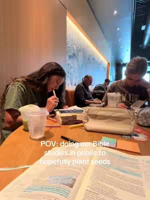 The image shows a brightly lit interior scene, likely a coffee shop. Two young women, dressed in casual attire, are seated at a wooden table. One is holding a straw near her mouth while looking at a book, and the other is reading. In the background, there's a glimpse of other people, some focused on their own activities. Books, drinks, and personal items are scattered on the table. Text overlay at the bottom states, 'POV: doing our Bible studies in public to hopefully plant seeds'.