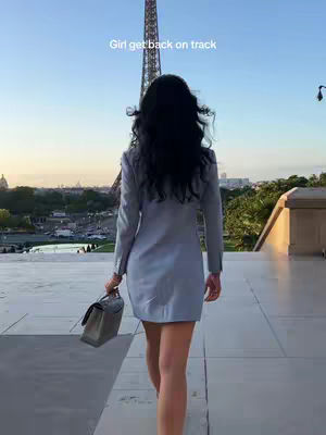 A woman with long dark hair, wearing a light blue dress, is walking away from the camera with the Eiffel Tower in the background. She is carrying a purse. The sky is clear and blue.