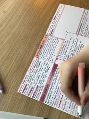 A top-down shot shows a notebook open on a wooden surface. The notebook pages are filled with handwritten notes, highlighted with pink and red markers. A hand holding a pink pen is visible, in the act of writing on the page which suggests a study environment. The background is a wooden table or desk.