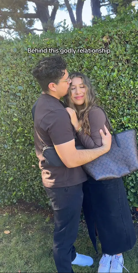 A couple is embracing in front of a green hedge. The man, wearing a brown shirt, glasses, and a crossbody bag, is kissing the woman's forehead. The woman, in a brown top and black skirt, carries a large handbag. The background is blurred, with bright sunlight.
