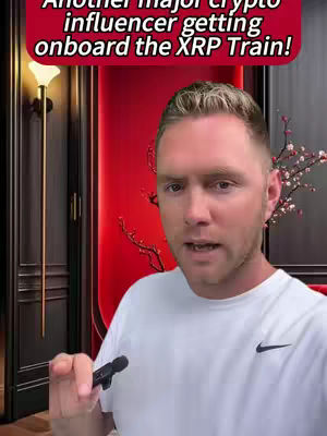 A man with fair skin and short blonde hair, wearing a white shirt with a small black logo, is speaking directly to the camera with a neutral expression. The background includes a red wall and what appears to be a dark wooden door and some type of floral decorations.