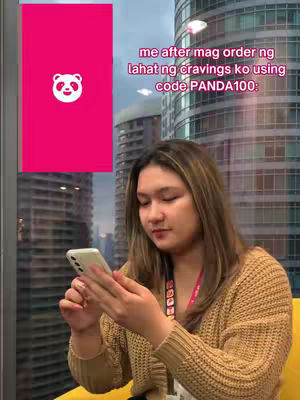 A young woman with long brown hair is seated near a window, using a smartphone. Behind her is a cityscape visible through the window, with tall buildings. A food delivery app logo, a panda, is on the left side of the frame, in front of a pink shape. Text overlay above the woman.