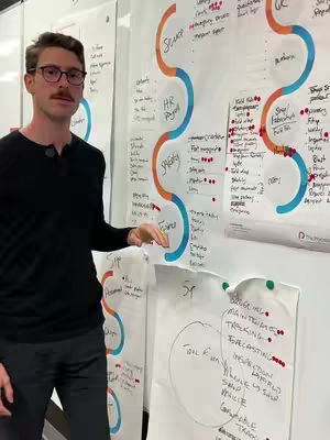 A man wearing glasses and a black shirt stands in front of various whiteboards. The whiteboards have color-coded diagrams and handwritten notes. The man is pointing towards one of the diagrams with his right hand.