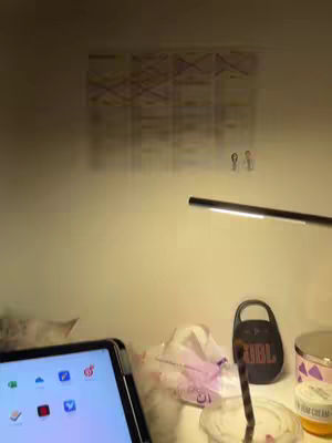 The image shows a well-lit workspace with a digital tablet in the foreground displaying app icons. Behind it, there's a tissue box, a water cup with a straw, and a small container of cream cheese. On the wall, a chart is pinned. A desk lamp provides light, casting shadows. The scene suggests a study or work setup, focusing on the tablet and surroundings.