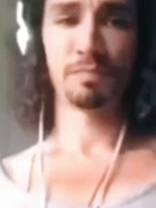 A close-up, slightly blurry image of a man's face. He has dark hair, a mustache, and a goatee. The man is wearing headphones and appears to be looking downcast. The lighting is subdued, creating a somewhat somber mood.