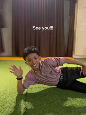 A young man is lying on a green surface, likely a floor, with his left arm propped up and his right arm extended. He wears a pink shirt and dark pants. The background features a brown curtain. The text overlay "See you!!!" is positioned above the man.