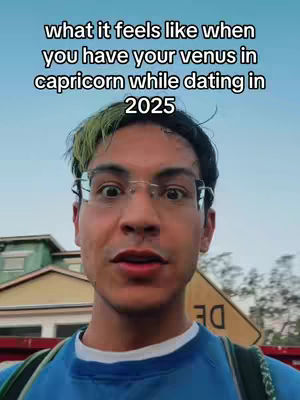 The cover image is a selfie-style shot featuring a young man with dark hair, styled with a touch of green dye on the bangs, wearing glasses and a blue shirt. His expression is open-mouthed, suggesting he's speaking or reacting to something. The background has a light blue sky and glimpses of a building and a road sign with the number 30 visible. The overlay text at the top says, 'what it feels like when you have your venus in capricorn while dating in 2025.' The overall composition is simple and direct, focusing on the creator's face with a clear, light backdrop.