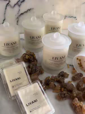 The image presents a collection of glass jars and wax melts, likely candles and wax. The jars are uniformly shaped, with clear glass bodies and white lids, some appear to be frosted. Each jar prominently displays the word 'LIVANI'. Surrounding the jars are clusters or loose pieces of what appears to be frankincense or a similar aromatic resin, also some wax melts. The products are arranged on a marble patterned surface. The overall composition is clean, well-lit, and designed to highlight the product.