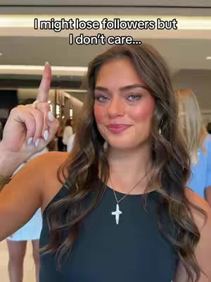 A woman with long brown wavy hair is front and center. She is wearing a black sleeveless top and a silver cross necklace, with a bright smile, pointing her index finger up, the background is slightly blurred in the background.
