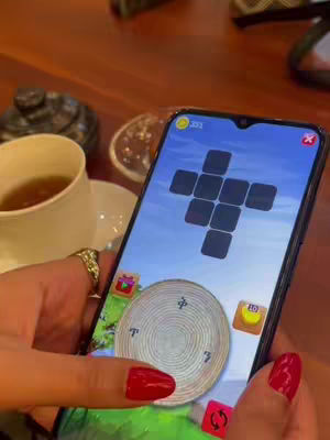 A hand with red nail polish holds a smartphone displaying a word puzzle game, with a teacup, saucer, and small dish visible in the background on a wooden table.