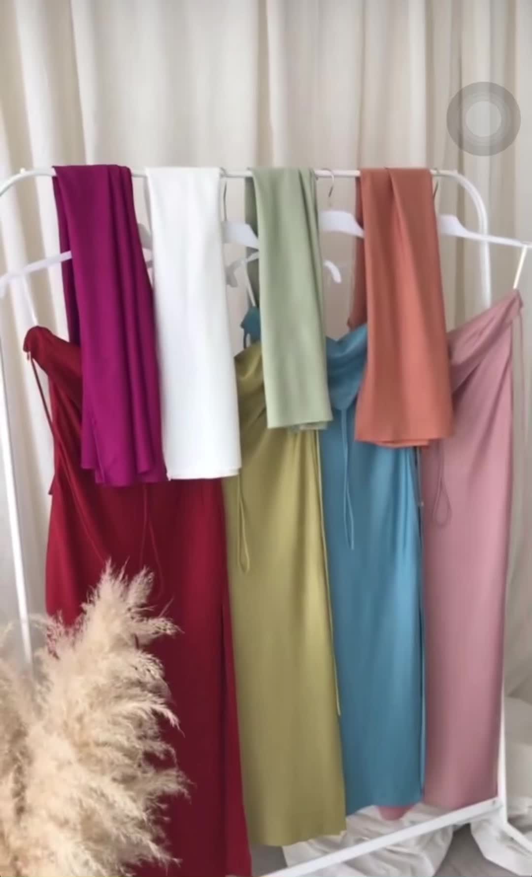 The image showcases a rack of dresses and fabric, each in a different solid color, hanging on a white rack. The dresses are visible at the bottom and various fabrics hang above them. In the foreground, there's a cluster of fluffy beige decorative grass.