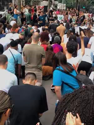 A medium shot of a crowd of people in an outdoor setting, likely a public space like a street. The focus is on a gathering of individuals, many of whom appear to be kneeling or bowing their heads. The image is saturated with natural light, and the perspective is from slightly above, looking down into the crowd, offering a wide view of the scene.