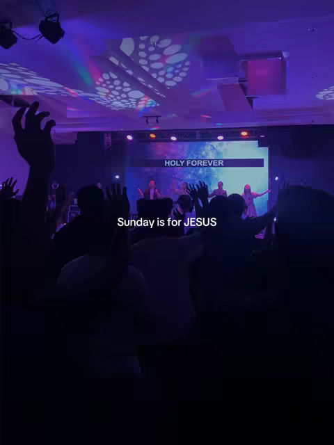 A photo from behind of a church service with many people raising their hands in worship. A stage is visible in the distance with words projected on a screen. The overall image is dark with a purple hue.