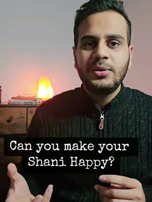 A man is centered in the frame, looking at the viewer with a serious expression. He is wearing a dark textured sweater. A text overlay highlights the question "Can you make your Shani Happy?" The background is simple with a chest of drawers to the left and a glowing lamp.