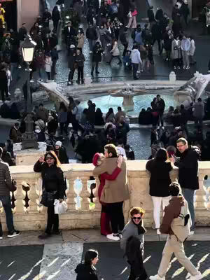 A bustling outdoor scene in Rome, Italy. The image is dominated by a crowd of people, with a focus on a couple embracing warmly in the middle ground. In the background, there's a fountain with other people present. The foreground is a stone structure.