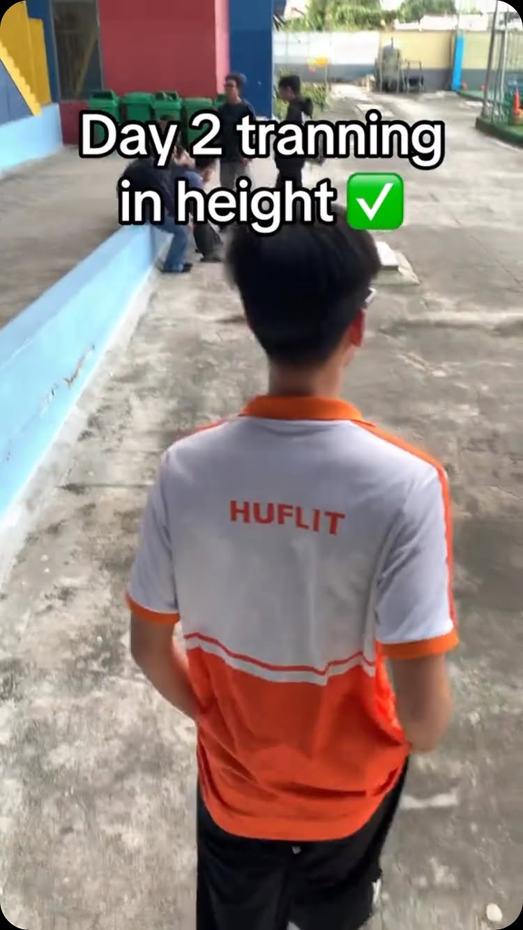 A shot from behind of a young man wearing a white and orange shirt, walking on a gray concrete surface. The text overlay reads 'Day 2 tranning in height' with a green check mark. Other people are visible in the background.