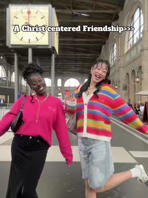 Two young women stand close together in front of a clock tower in what appears to be a European city. The woman on the left is wearing a pink sweater and a black skirt, while the woman on the right is wearing a colorful striped cardigan, denim shorts, and sneakers. The text overlay reads, "A Christ centered Friendship>>>".