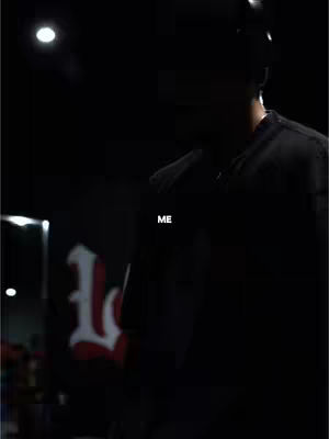 A dimly lit scene featuring a person in a dark-colored shirt standing in front of what appears to be a stage or performance area. The person is silhouetted, making their features indistinct. The word 'ME' is overlaid on the image in white, and a logo with a large letter 'L' is partially visible in the background.