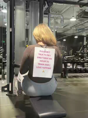 A person, viewed from the back, sits on a gym bench in an active gym environment. A sheet of paper is pinned to their back with text, providing a humorous element. The setting incorporates gym equipment and ambient lighting, suggesting a workout context.