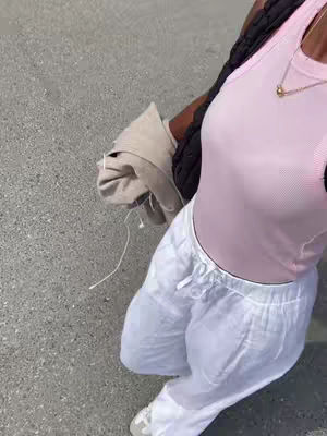 A person in casual clothing, primarily in pink and white, stands on a paved surface, viewed from a slightly elevated angle. The person is seen from the torso upwards, with emphasis on the upper body. The individual wears a light pink tank top, white linen pants, and sneakers. They seems to be wearing a black structured item over one shoulder. The background is a grey asphalt-like surface.
