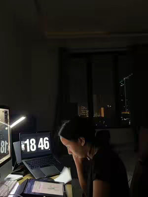 A dimly lit room features a person seated, illuminated by the glow of a computer and a laptop. The scene conveys a sense of late-night studying or work, with city lights visible through a window in the background.