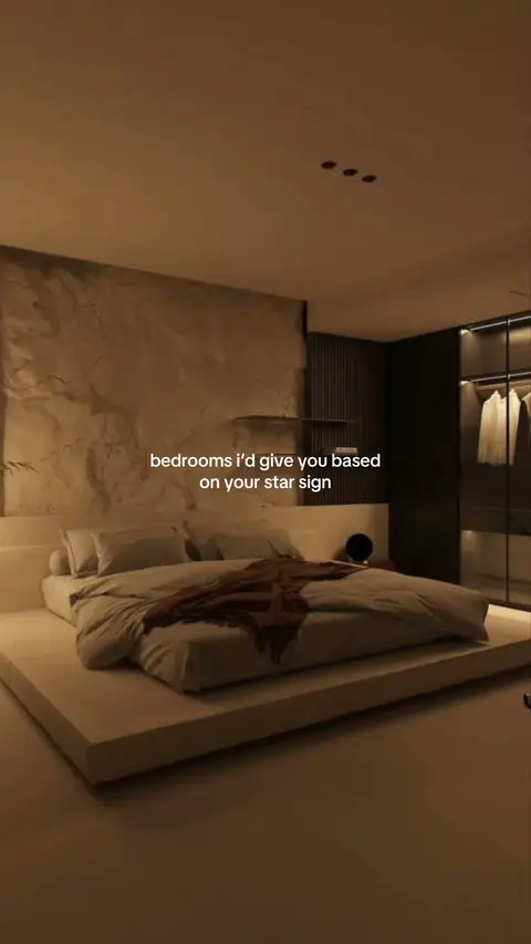 A dimly lit interior shot of a modern bedroom featuring a large, minimalist platform bed covered with neutral-colored bedding and a brown throw blanket. The backdrop is a textured wall with a large headboard. To the right, there's a closet with hanging clothes visible through a glass door. The room is bathed in warm, ambient lighting.