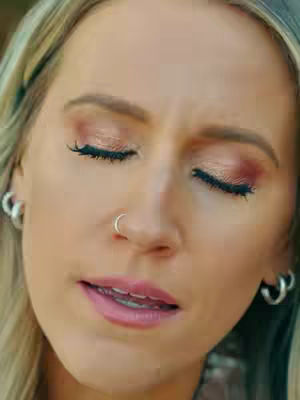 A close-up shot features a woman with blonde hair and soft makeup, including pink eyeshadow and lip color. Her eyes are closed, and she has a subtle smile. She wears silver hoop earrings and a silver nose ring.