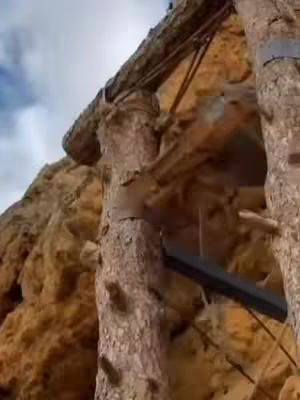 An upward-angled shot of what appears to be a wooden structure built into a rock face. The foreground features the rough, textured surface of the structure, made from what looks like timber. The color palette is dominated by shades of brown and tan, with the sky peeking through as a light blue at the top.