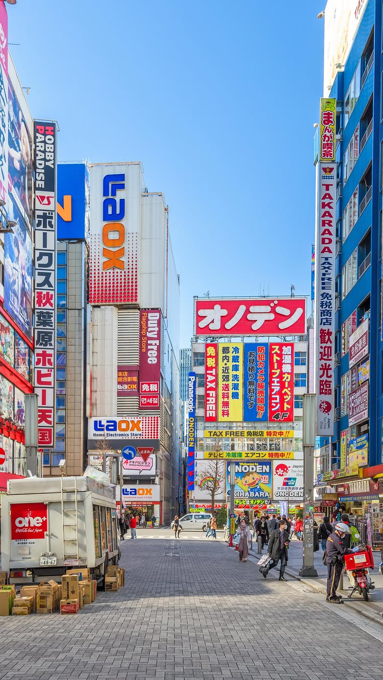 A wide, daytime shot of a crowded city street in Tokyo, Japan. Tall buildings with colorful signs and advertisements line both sides of the street, creating a narrow, urban canyon effect. The sky is a clear, vibrant blue. People are walking along the sidewalks and in the street, and a delivery truck and parked vehicles are visible in the foreground. The overall feel is bustling and energetic.