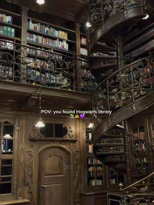 An interior shot of a grand library with wooden paneling, ornate spiral staircase, and towering bookshelves filled with books. Overhead, there are several hanging elegant light fixtures.