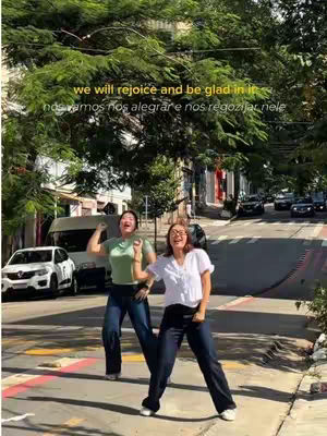Two women dancing joyfully in a street scene, arms raised, under a canopy of green trees and a sky obscured by foliage. The scene is vibrant with natural light, featuring a background of buildings, a road, and vehicles, suggesting an urban setting. Text overlays their dancing figures, enhancing the jovial essence of the moment.