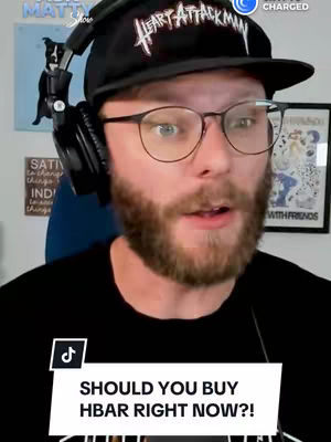 A man with glasses and a beard is in the foreground, wearing a black cap that says 'Heart Attack Man' and a black t-shirt. He is wearing headphones. In the background are items on a wall, including a painting of a dog wearing headphones. A white rectangle at the bottom of the frame has the text 'SHOULD YOU BUY HBAR RIGHT NOW?!'.