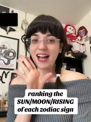 A woman with dark hair and glasses is smiling and holding out her left hand toward the viewer, in front of a background of a wall with framed art and a shelf with dolls. The woman is facing the camera, with open eyes and mouth. The image is well-lit, and the background is slightly blurred. A text overlay sits at the bottom.