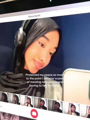 The image is a selfie taken on a computer screen. The main subject is a young woman wearing a black hijab and headphones. She is looking slightly to the side with a soft smile. A text overlay in white font is present on the image, and the background is a muted blue. Below the main image are smaller thumbnails of the woman in different poses, suggesting a photo booth or similar interface.
