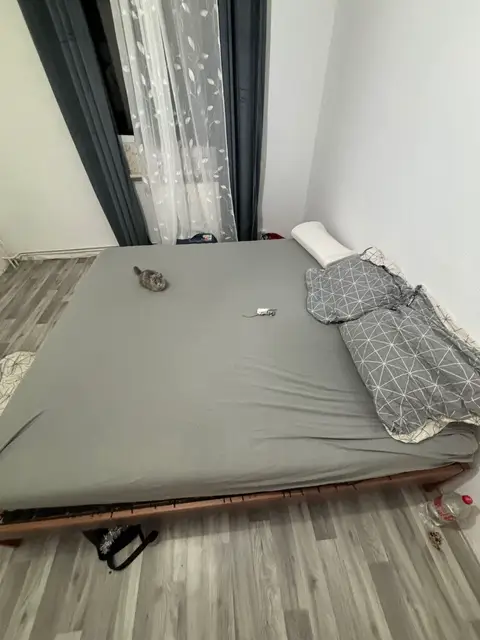 An overhead shot of a bed in a bedroom. The bed has a gray sheet, a white pillow, and a blanket with a geometric pattern. The floor is light-colored wood. A small, soft toy is on the sheet. The room has a window with curtains.