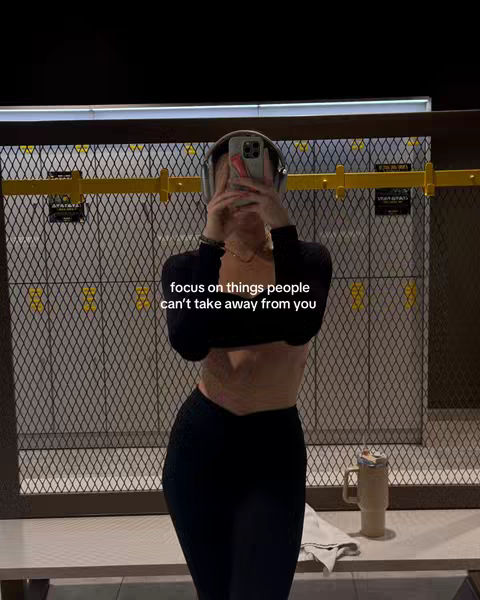 A selfie-style image taken in what appears to be a gym locker room. The person is holding up a phone to cover their face. The background features metal lockers, yellow supports, and a white bench with a towel. The overall lighting is neutral.