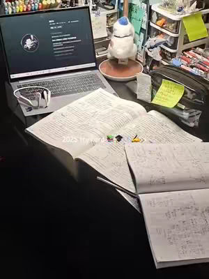 A desk with a laptop, books, and study materials. The laptop screen displays a profile page. Open books are spread out, with notes and writing visible. Headphones sit near the laptop. A white decorative accessory and school supplies are in the background. The color palette is dominated by dark tones with highlights from the screen and books.