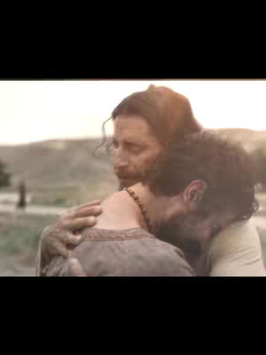 The image captures a tender moment between two people, likely in a historical or biblical setting. One person, likely representing Jesus, has their arms wrapped around another, who appears to be leaning against him. The scene is bathed in soft, warm light, with a blurred background of a field or landscape. The overall composition evokes feelings of comfort, protection, and compassion.