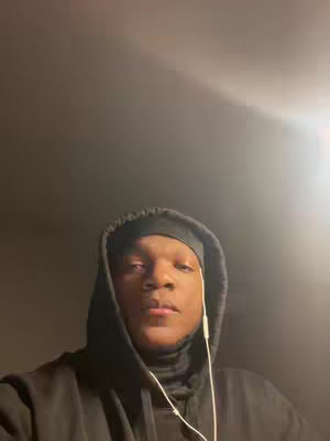 A medium shot shows a young person with a dark complexion centered in the frame. They wear a black hoodie with the hood up and a black cap underneath, obscuring most of their hair. White earbuds dangle, and a dark background surrounds them. The lighting is soft and comes from above.