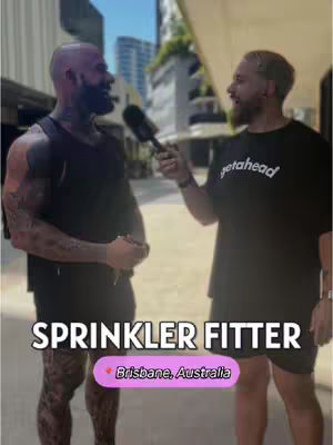 Two muscular men are standing in front of a building in Brisbane, Australia. One man is holding a microphone and the other is the subject of the interview. The text overlay reads "SPRINKLER FITTER" with a pink bubble containing"Brisbane, Australia".