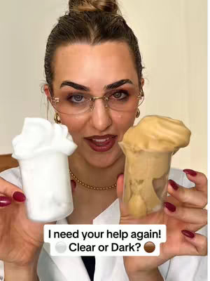 A woman with light skin and brown hair styled in a topknot holds two transparent cups filled with a foamy substance. The left cup contains a white substance, while the right cup contains a tan substance. She wears eyeglasses with gold rims, a gold necklace, and rings on her fingers. Her nails are painted dark red. Text on the image asks for a choice between "Clear or Dark?" with corresponding circle icons.