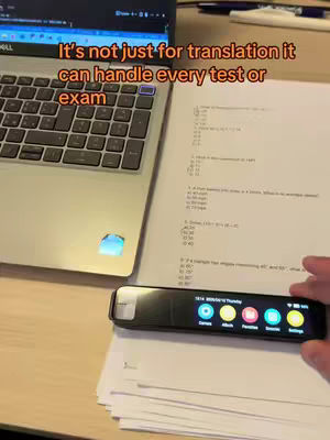 The image shows a laptop, a sheet of paper with a test on it, and a handheld device. The laptop is on the left, open, and reflecting light. The sheet of paper with the test lies on the desk to the right, and the handheld device is in the bottom right. The handheld device has a screen showing icons.