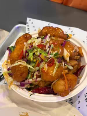 A close-up shot of a colorful meal served in a white bowl. The bowl contains a mix of cooked food items, including what appears to be fried dumplings with a glossy sauce, mixed with vegetables and red chili peppers. The food rests on a white paper with printed text. The image is shot from a slightly high angle, giving a clear view of the dish.