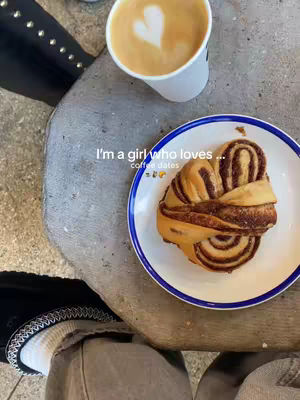 The cover image shows a top-down view of someone's legs and a table. On the table, there's a plate with a pastry (likely a cinnamon roll) and a cup of coffee with a heart-shaped design in the foam is placed. The composition suggests a cozy, brunch-like setting. The legs are clad in a pair of jean-like pants with white socks and shoes. The table has rough, stone-like texture with added text that says "I'm a girl who loves...coffee dates".