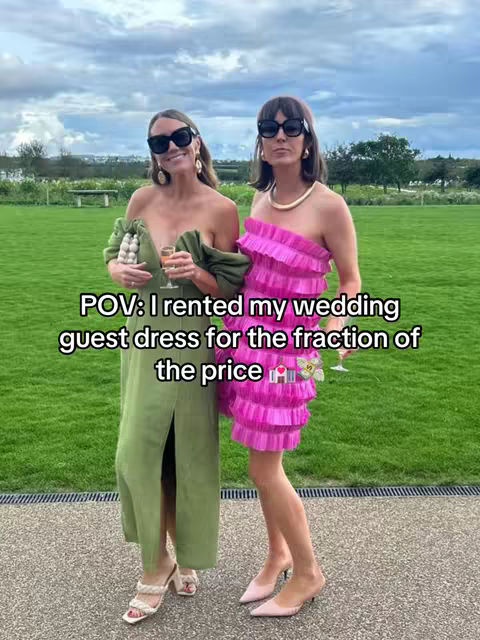 Two women are standing side-by-side in front of a green grassy field, with a cloudy sky in the background. They are both wearing sunglasses and smiling, and appear to be at a wedding. The woman on the left is wearing a long green dress and holding a small purse and a glass of champagne. The woman on the right is wearing a pink ruffled dress with a gold necklace.